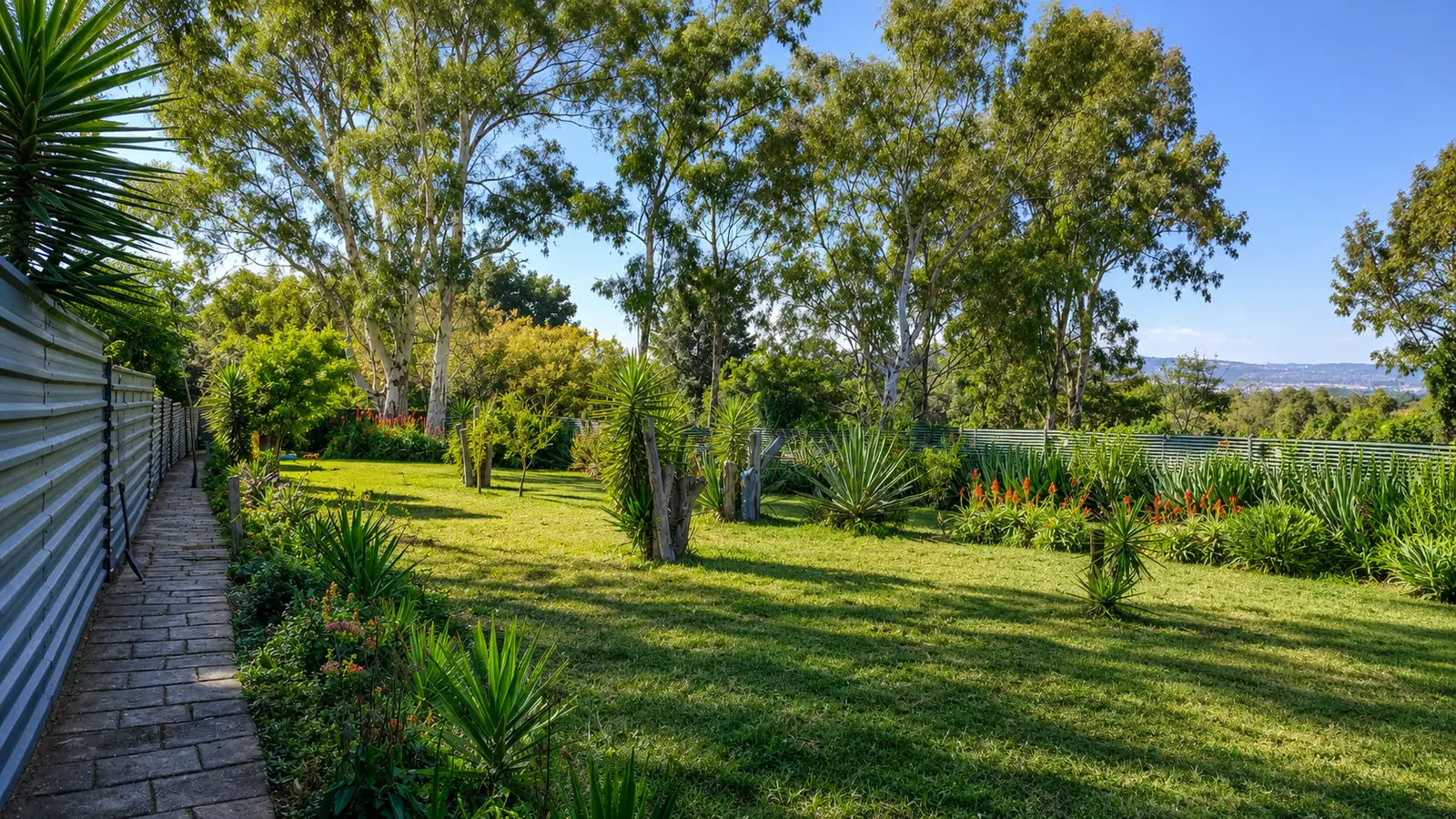 Landscaped estate garden — a paved path along the perimeter fence, mature gum trees, lawns and flowering aloes under a clear Highveld sky